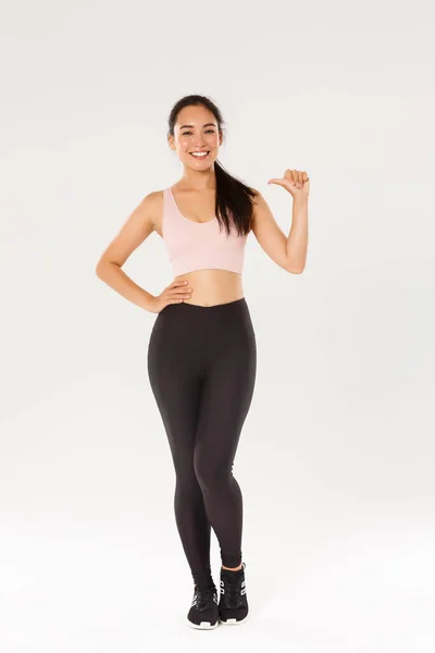 Comprimento total de confiante sorrindo asiática atleta feminina em sportsbra e leggigns, menina fitness apontando para si mesma, levar estilo de vida ativo e saudável, ser profissional, de pé fundo branco — Fotografia de Stock