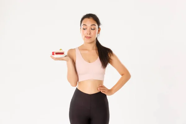 Estilo de vida ativo, fitness e conceito de bem-estar. Bonito morena asiático menina atleta, tentador para comer delicioso bolo doce, mas estar na dieta, cuidar do peso corporal e calorias, fundo branco — Fotografia de Stock