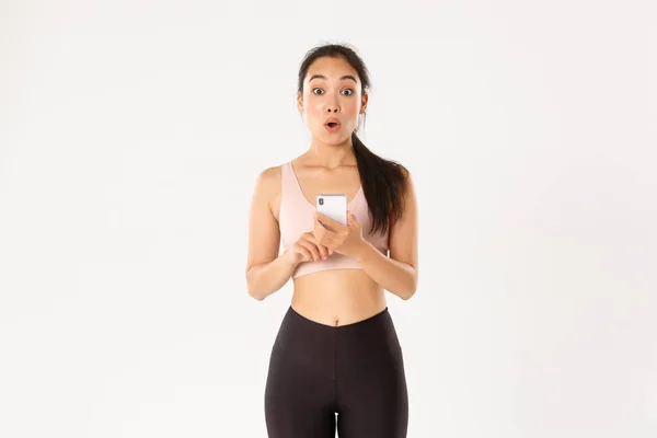 Sport, technology and active lifestyle concept. Surprised and amazed asian female athlete, sportswoman say wow, checking fitness app on mobile phone, using workout tracker, running with application — Stock Photo, Image