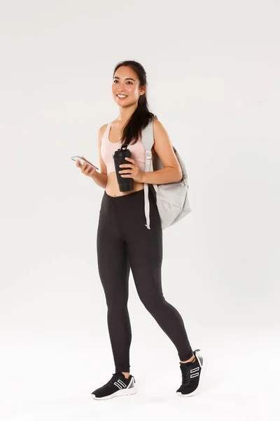 Longitud completa de sonreír sano, delgado asiático chica ir entrenamiento de fitness, hembra athelte llevar mochila con equipo de entrenamiento y botella de agua, el uso de la aplicación de deportes de teléfono móvil, fondo blanco — Foto de Stock