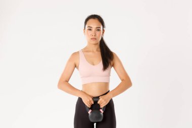 Spor, refah ve aktif yaşam tarzı konsepti. Ciddi ve odaklanmış Asyalı güçlü fitness kızı, spor salonunda bayan sporcu, kettlebell egzersizi, ayakta duran beyaz arka plan