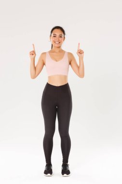 Spor, spor salonu ve sağlıklı vücut konsepti. Neşeli bir gülümseme ve hoş bir fitness kızı, parmaklarını havaya kaldırarak egzersiz malzemeleri reklamını gösteriyor, eğitim için üyelik satın alıyor.