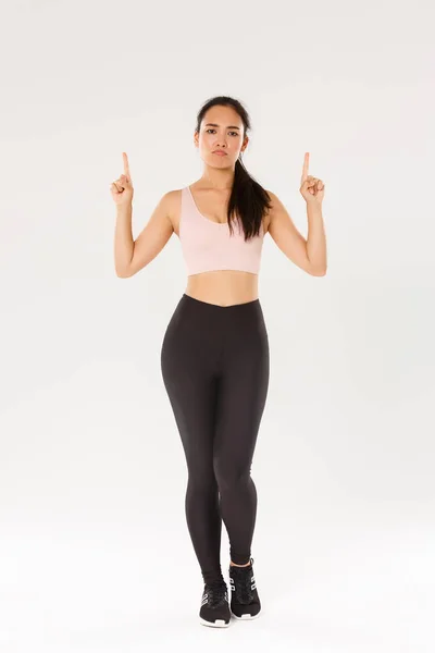 Sport, gymnase et concept de corps sain. Pleine longueur de fille de fitness non amusée et sceptique, athlète féminine asiatique en vêtements de sport se plaignant, boudant les doigts mécontents et pointant vers le haut, fond blanc — Photo