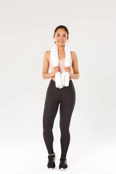 Esporte, ginásio e conceito de corpo saudável. Comprimento total de sorrir bonito menina slim, treinador de fitness ou esportista após exercícios no ginásio, de pé com toalha enrolado em torno do pescoço, fundo branco — Fotografia de Stock