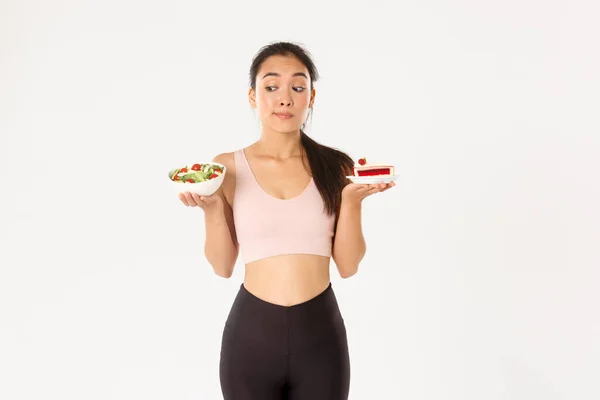 Активный образ жизни, фитнес и велнес. Нерешительная милая азиатская стройная девочка в спортивном костюме, держит здоровый салат и торт, колеблется, нужно оставаться на диете, похудеть, белый фон — стоковое фото