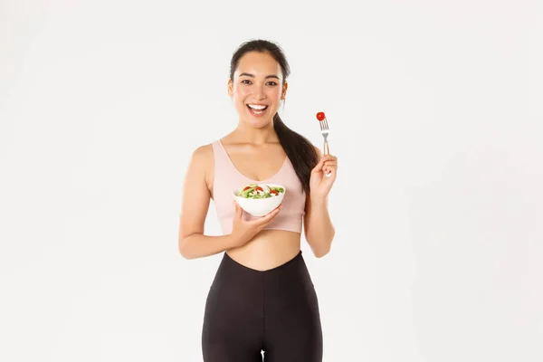 Esporte, bem-estar e conceito de estilo de vida ativo. Sorrindo magro e saudável menina asiática fazendo exercícios de fitness, perder peso com treino de ginásio e comer saladas frescas, de pé fundo branco — Fotografia de Stock