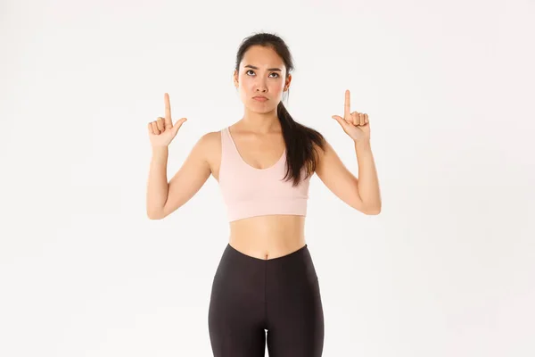 Deporte, bienestar y concepto de estilo de vida activo. Molesto y decepcionado deportista asiático, mujer en ropa de fitness señalando y mirando hacia arriba con cara de enfurruñamiento, quejándose en la pancarta, fondo blanco —  Fotos de Stock