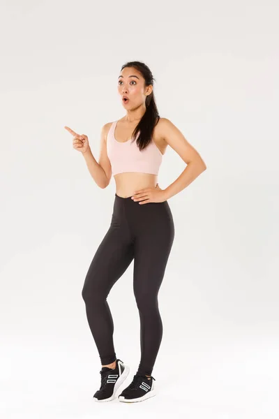 Concepto de deporte y gimnasio. Longitud completa de sorprendido y sorprendido linda chica asiática en ropa de fitness, señalando y mirando esquina superior izquierda impresionado, mostrando el equipo de entrenamiento o gimnasio precios de membresía — Foto de Stock