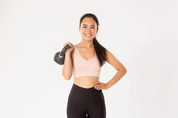 Esporte, bem-estar e conceito de estilo de vida ativo. Sorrindo forte e magro asiático fitness menina, atleta feminino segurando kettlebell e olhando despreocupado, ganhando músculos no ginásio, de pé fundo branco — Fotografia de Stock