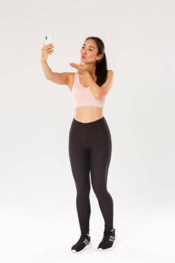 Spor kıyafetleri giyip spor salonundan selfie çeken ya da spor salonundan canlı yayın yapan, cep telefonu ekranına hava öpücüğü gönderen, beyaz arka planda duran, spor salonundaki sevimli ve aptal kadın blogcu.