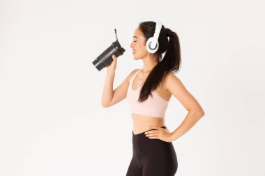 Spor, refah ve aktif yaşam tarzı konsepti. Tatmin olmuş gülümseme ve kulaklıklı tatlı Asyalı kız profili, antreman sırasında su molası, müzik eşliğinde verimli bir eğitim.
