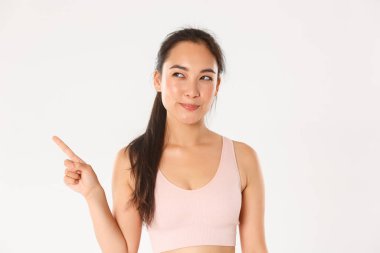 Spor, refah ve aktif yaşam tarzı konsepti. Kurnaz, düşünceli, gülümseyen Asyalı sporcu, seçimini yapan fitness kızı, meraklı bir gülümsemeyle mutlu görünüyor, sol üst köşeyi işaret ediyor.