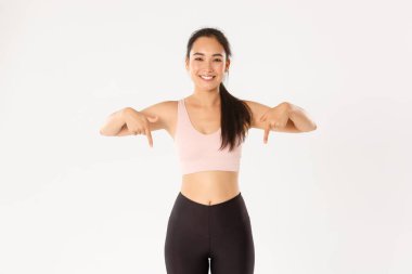Spor, refah ve aktif yaşam tarzı konsepti. Gülümseyen ve zayıf kadın fitness eğitmeni, spor salonunda çalışan Asyalı kız ve bir şey gösteren, parmaklarını yere doğru gösteren, beyaz arka planda duran