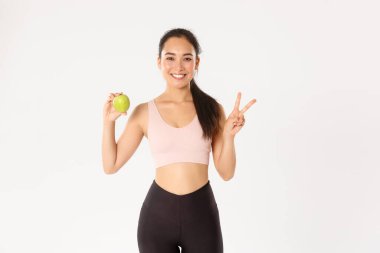 Spor, refah ve aktif yaşam tarzı konsepti. Çekici Asyalı kadın fitness koçu, kız antrenör tavsiyesi egzersiz ve antrenmandan sonra sağlıklı yemek yeme, elmayla ayakta durma, barış işareti göster
