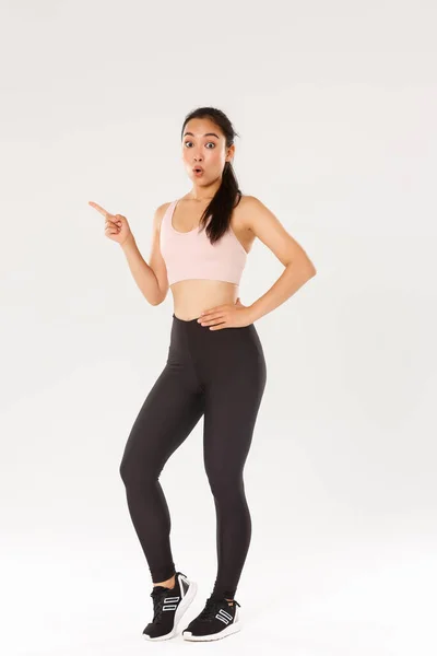 Deporte, gimnasio y concepto corporal saludable. longitud completa de sorprendido lindo morena asiática chica de fitness en ropa deportiva, mirando sorprendido, señalando el dedo a la izquierda, mostrando precios impresionantes para el equipo de gimnasio —  Fotos de Stock