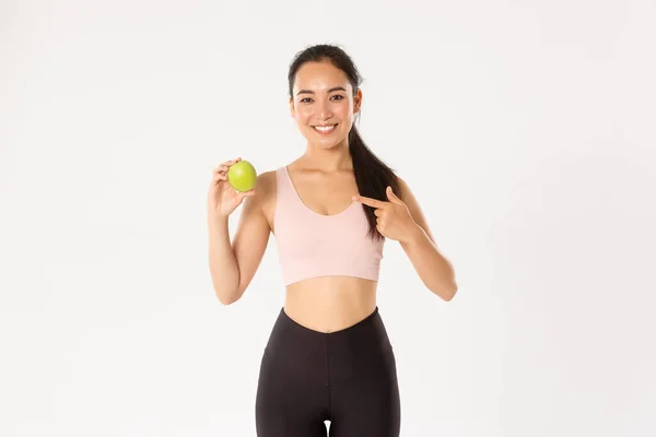 Esporte, bem-estar e conceito de estilo de vida ativo. Retrato de sorrir magro e ajuste asiático fitness girl, treino treinador conselhos comer vitaminas e alimentos saudáveis, apontando para maçã verde, fundo branco — Fotografia de Stock