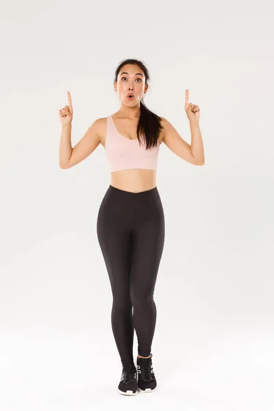 Longitud completa de atelta femenina asiática sorprendida e impresionada, deportista en ropa deportiva mirando y señalando con los dedos hacia arriba con la cara excitada. Chica fitness se preguntó con oferta especial en tienda de deportes — Foto de Stock