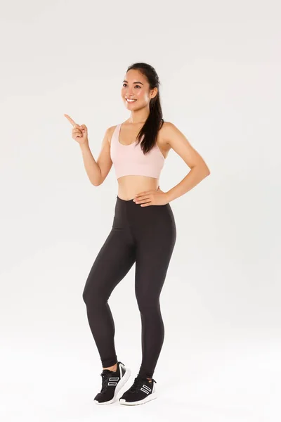 Deporte, gimnasio y concepto corporal saludable. Longitud completa sonriendo atleta femenina satisfecha, chica asiática fitness en desgaste activo haciendo su elección, señalando y mirando esquina superior izquierda, recoger gimnasio para el entrenamiento — Foto de Stock