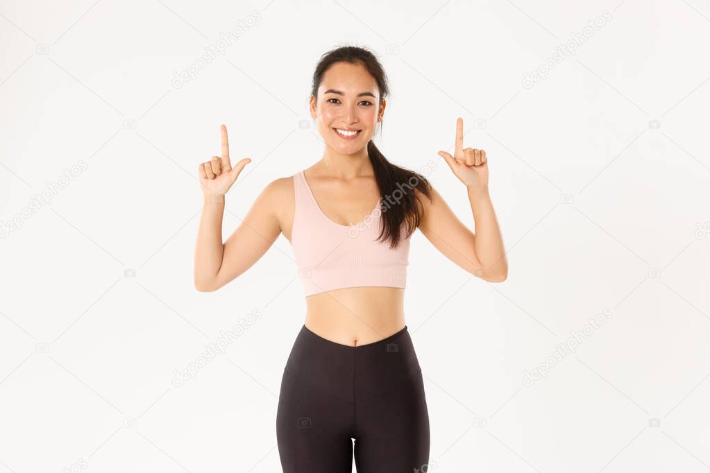 Sport, wellbeing and active lifestyle concept. Smiling slim and cute asian fitness girl, female athlete pointing fingers up, showing workout equipment sale promo or gym membership offer.