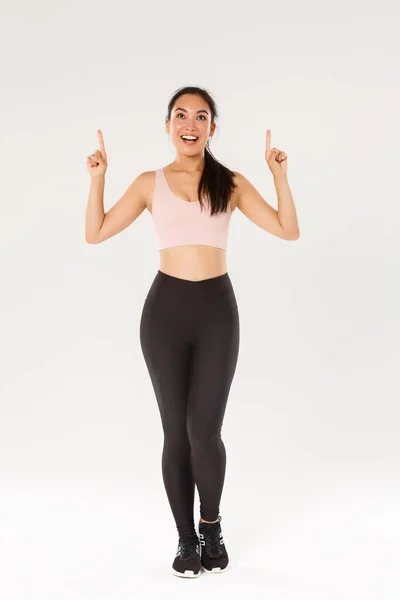 Esporte, ginásio e conceito de corpo saudável. Comprimento total do atleta feminino sorridente animado em sportswear olhando espantado e apontando os dedos para cima, atleta feminino desfrutando de fitness, convidar novos membros — Fotografia de Stock