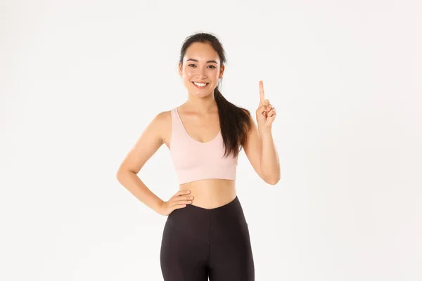 Esporte, bem-estar e conceito de estilo de vida ativo. Sorrindo asiático instrutor de fitness, menina em sportswear ensino lição, mostrando regra número um, levantar o dedo e sorrir, pé fundo branco — Fotografia de Stock