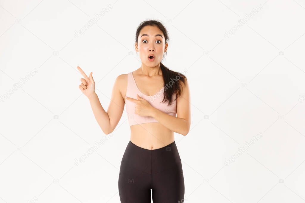 Sport, wellbeing and active lifestyle concept. Impressed and excited sportswoman, asian athlete in activwear open mouth wondered and pointing fingers upper left corner at your logo banner.