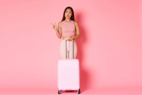 Reisen, Urlaub und Urlaubskonzept. Ganzes zweifelhaftes und verwirrtes asiatisches Touristenmädchen, das zögerlich nach links zeigt und mit einem Koffer unentschlossen vor rosa Hintergrund steht — Stockfoto