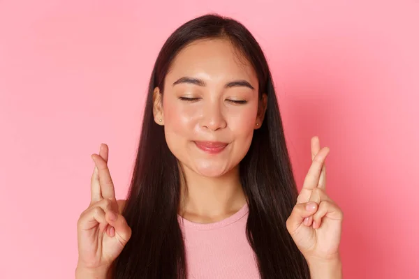 Skønhed, mode og livsstil koncept. Close-up af håbefuld optimistisk asiatisk pige, der ønsker, lukker øjnene og smiler afslappet, mens krydser fingre held og lykke og beder drøm går i opfyldelse, lyserød baggrund - Stock-foto