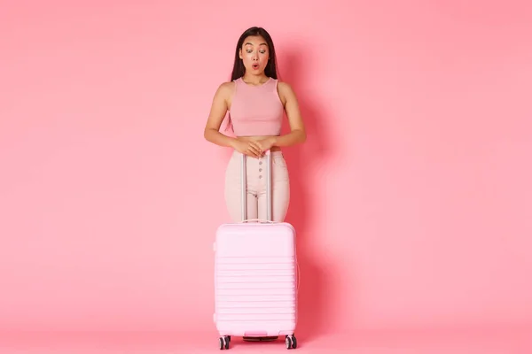 Viajar, férias e férias conceito. Comprimento completo de divertido e surpreso asiático menina bonita em roupas de verão olhando espantado com mala, olhar para baixo, de pé fundo rosa — Fotografia de Stock