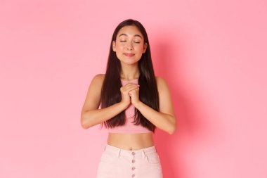 Yaşam tarzı, güzellik ve kadın konsepti. Rüya gibi güzel Asyalı bir kızın portresi, gözlerini kapat ve gülümse ellerini birbirine bağlayıp dua ederken, yalvarırken, dua ederken, Tanrı 'dan dilek dilerken, pembe arka plan