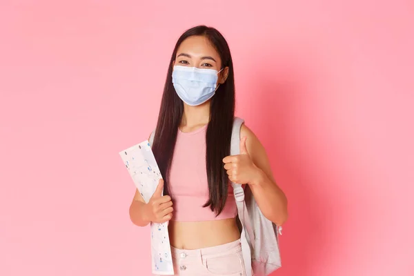 Güvenli turizm, koronavirüs salgını sırasında seyahat etmek ve virüs konseptini önlemek. Neşeli, sevimli Asyalı turist kız yurt dışına seyahat ederken sosyal mesafeyi teşvik ediyor, baş parmağını kaldırıyor ve tıbbi maske takıyor. — Stok fotoğraf
