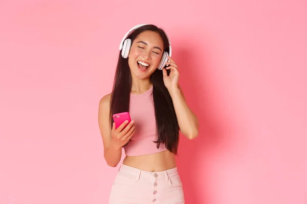 ヘッドフォンで音楽を聴きながら、携帯電話でカラオケゲームをプレイし、ピンクの背景の上に立って、魅力的なスタイリッシュなアジアの女の子の近くの目と歌に沿って歌うのウエストアップの肖像画 — ストック写真