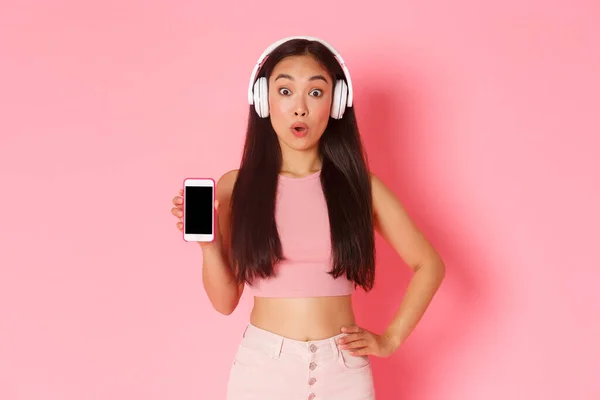 Technologia, komunikacja i koncepcja stylu życia online. Imponująca azjatycka dziewczyna znalazła niesamowite podcast, pokazując ekran smartfona jak noszenie słuchawek, słuchanie muzyki, różowe tło — Zdjęcie stockowe