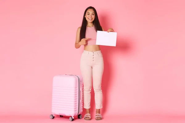 Viaggiare, vacanze e concetto di vacanza. Full-length di sorridente attraente ragazza asiatica in attesa di qualcuno in aeroporto, puntando il dito verso un pezzo di carta bianco con valigia su sfondo rosa — Foto Stock