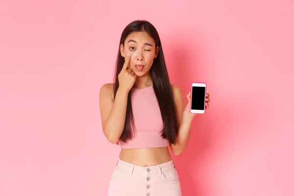 Tecnologia, comunicação e conceito de estilo de vida online. Silly menina asiática puxando pálpebra e mostrando tela do smartphone, avisar namorado como ela olhando sobre sua atividade de mídia social, zombando pessoa — Fotografia de Stock