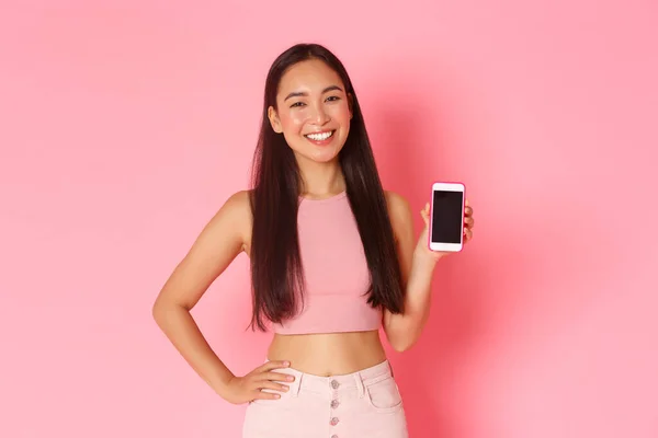 Technologie, komunikace a koncepce on-line životního stylu. Nádherný trendy asijské dívka stojící nad růžovým pozadím s mobilním telefonem, zobrazování app nebo smartphone obrazovka banner, růžové pozadí — Stock fotografie