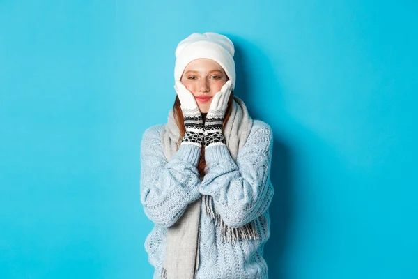 겨울 과 휴일 개념. 하얀 비니를 입고 스웨터를 입고 장갑을 끼고 볼을 꽉 쥐고 뿌듯 한 미소를 머금은 귀여운 소녀가 푸른 배경 위에 서서 — 스톡 사진