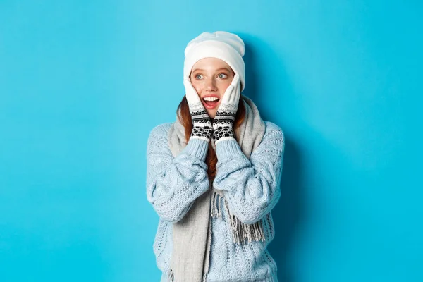 겨울 옷, 비니, 장갑, 스웨터를 입고 파란 배경 위에 서서 로고를 보고 흥분 한 빨간 머리 소녀 — 스톡 사진