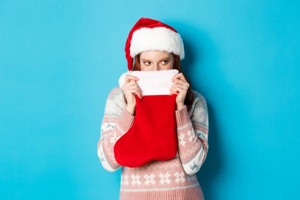 Bonito rosto tampa menina com meia de Natal, olhando para a direita com olhar astuto, de pé em chapéu de Santa e celebrando férias de inverno, fundo azul — Fotografia de Stock