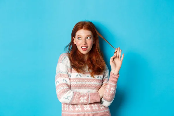 Flirty redhead girl staring left, playing with hair strand and thinking, standing over blue background