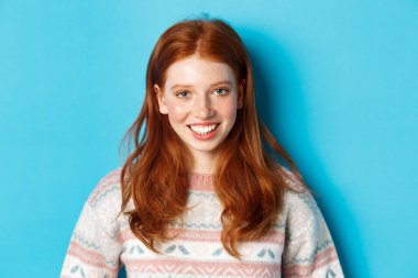Süveter giyen, kameraya mutlu gülümseyen, mavi arka planda duran tatlı kızıl saçlı kızın yakın çekimi.