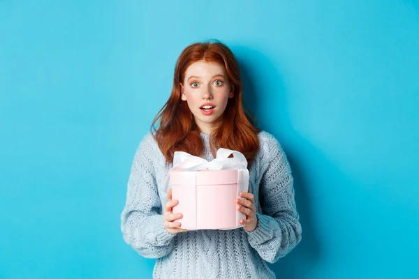 Surprised redhead girl receiving valentines gift, holding box with present and staring at camera amazed, wearing sweater, standing over blue background — Stock Photo, Image