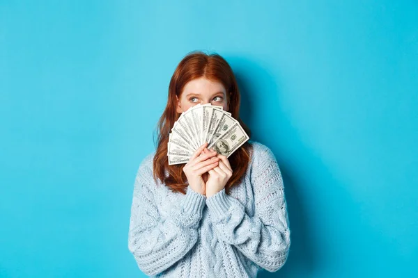 Chica linda pensativa con el pelo rojo soñando con ir de compras, sosteniendo dólares y mirando el logotipo de la esquina superior izquierda, de pie sobre el fondo azul — Foto de Stock