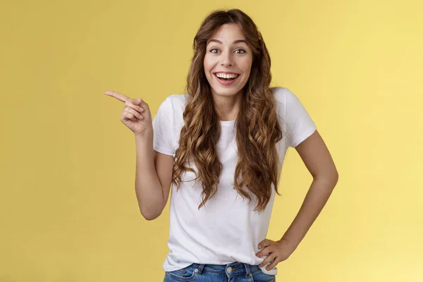 Animado surpreendido feliz entusiasta boa aparência mulher longo encaracolado penteado branco t-shirt rindo impressionado surpreso apontando dedo indicador esquerdo discutir interessante exposição amarelo fundo — Fotografia de Stock