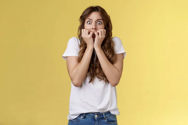 Scioccato senza parole spaventato stordito dai capelli ricci donna caucasica ansimante tenere le dita bocca mordere chiodi fissare fotocamera spaventato esprimere paura terrorizzato spaventoso film stand sfondo giallo — Foto Stock