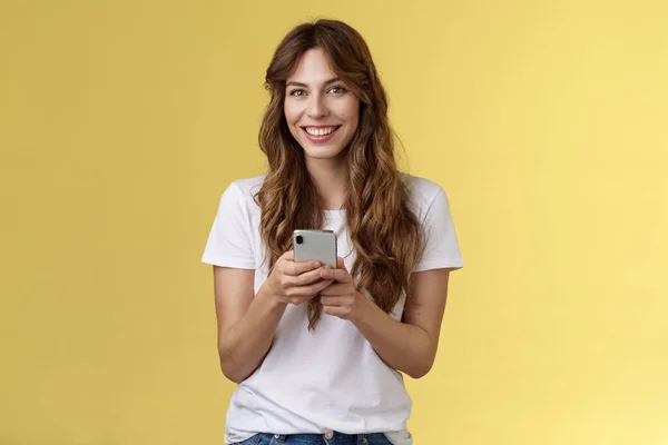 Entusiastiska charmiga sällskaplig ung flicka meddelande vän skicka foton sociala medier håll smartphone ser ut kamera glatt vänlig leende stå gul bakgrund casual outfit — Stockfoto