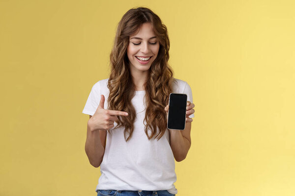 Intrigued charismatic happy young female blogger show what photo app use edit social media profile hold smartphone looking pointing index finger mobile phone blank screen smiling amused