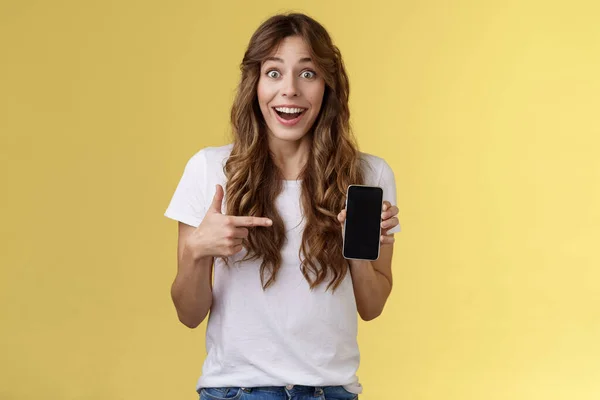 Surprised happy lucky girl winning online internet lottery smiling broadly hold smartphone pointing blank mobile phone screen showing display grinning excited enthusiastic stand yellow background — Stock Photo, Image