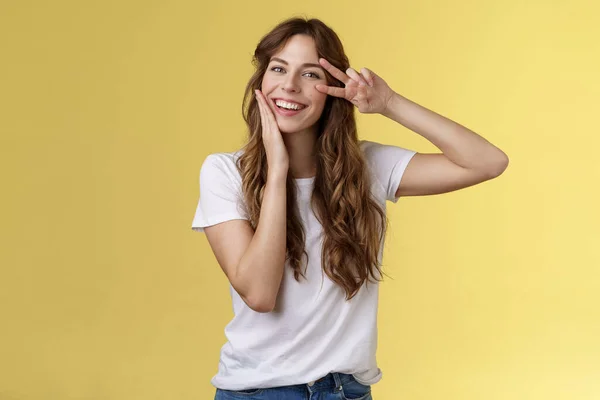 Cheerful positive lovely girlfriend curly hairstyle touch cheek blushing modest cute showing victory peace sign near eye optimistic upbeat attitude having fun enjoying summer yellow background — Stock Photo, Image