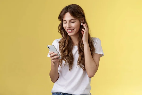 Tender feminine curly-haired female white t-shirt touch wireless earbud put earphone ear smiling pleased look smartphone screen pick song want to listen music search right track yellow background — Stock fotografie